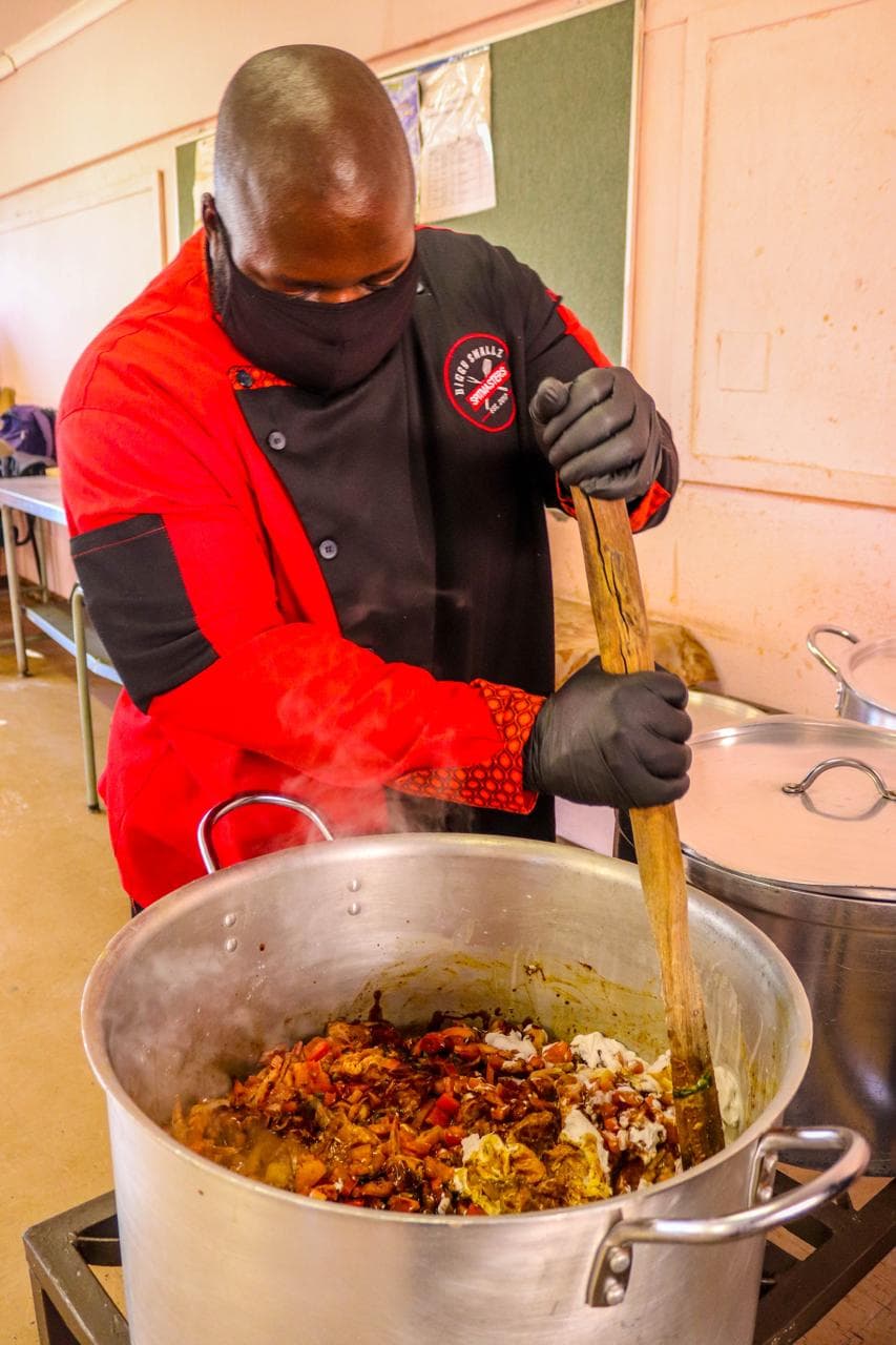 Biggy stirring a large pot of stew over the heat, masked and gloved.