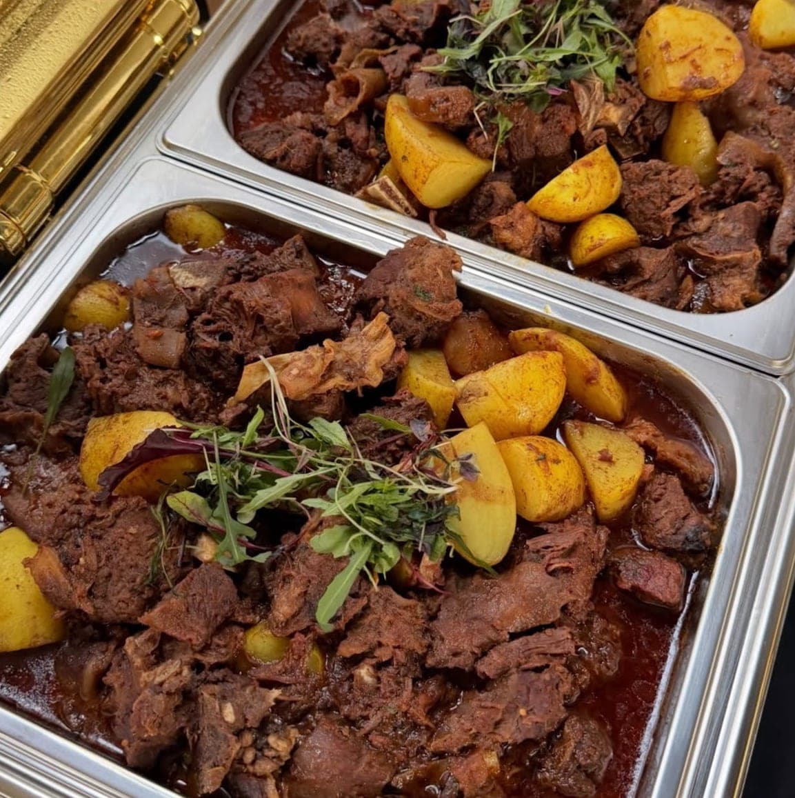 Two serving trays of Beef Rendang Curry with potatoes, topped with fresh leaves.