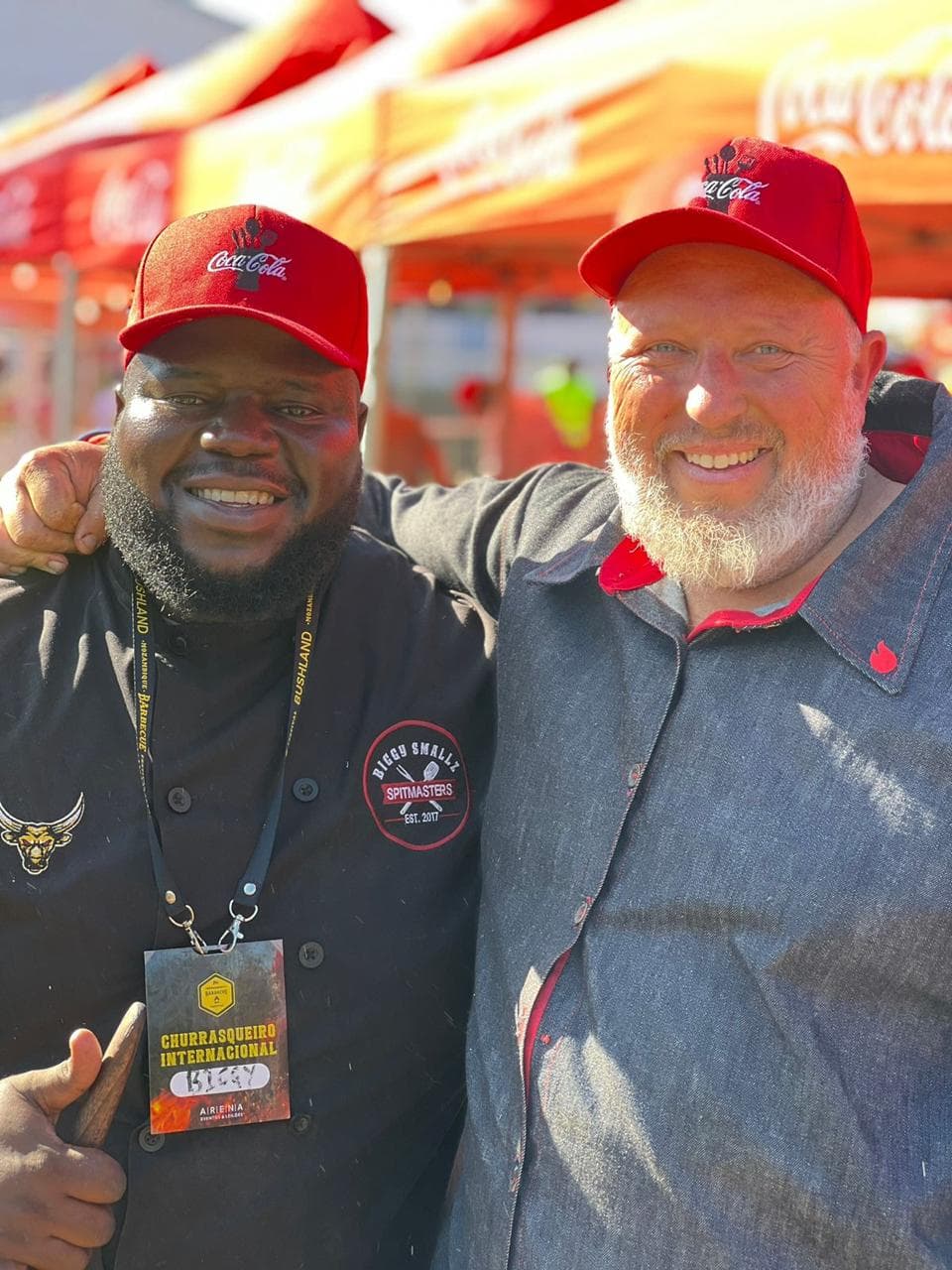 Biggy and a fellow chef at the festival, both in Coca-Cola caps, the Churrasqueiro Internacional lanyard around Biggy's neck.