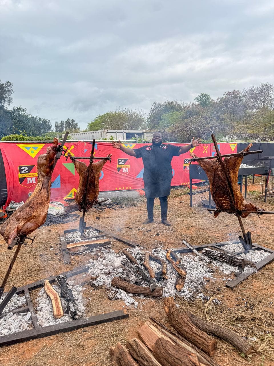Lamb halves roasting on stands beside a fire, cloudy sky overhead.