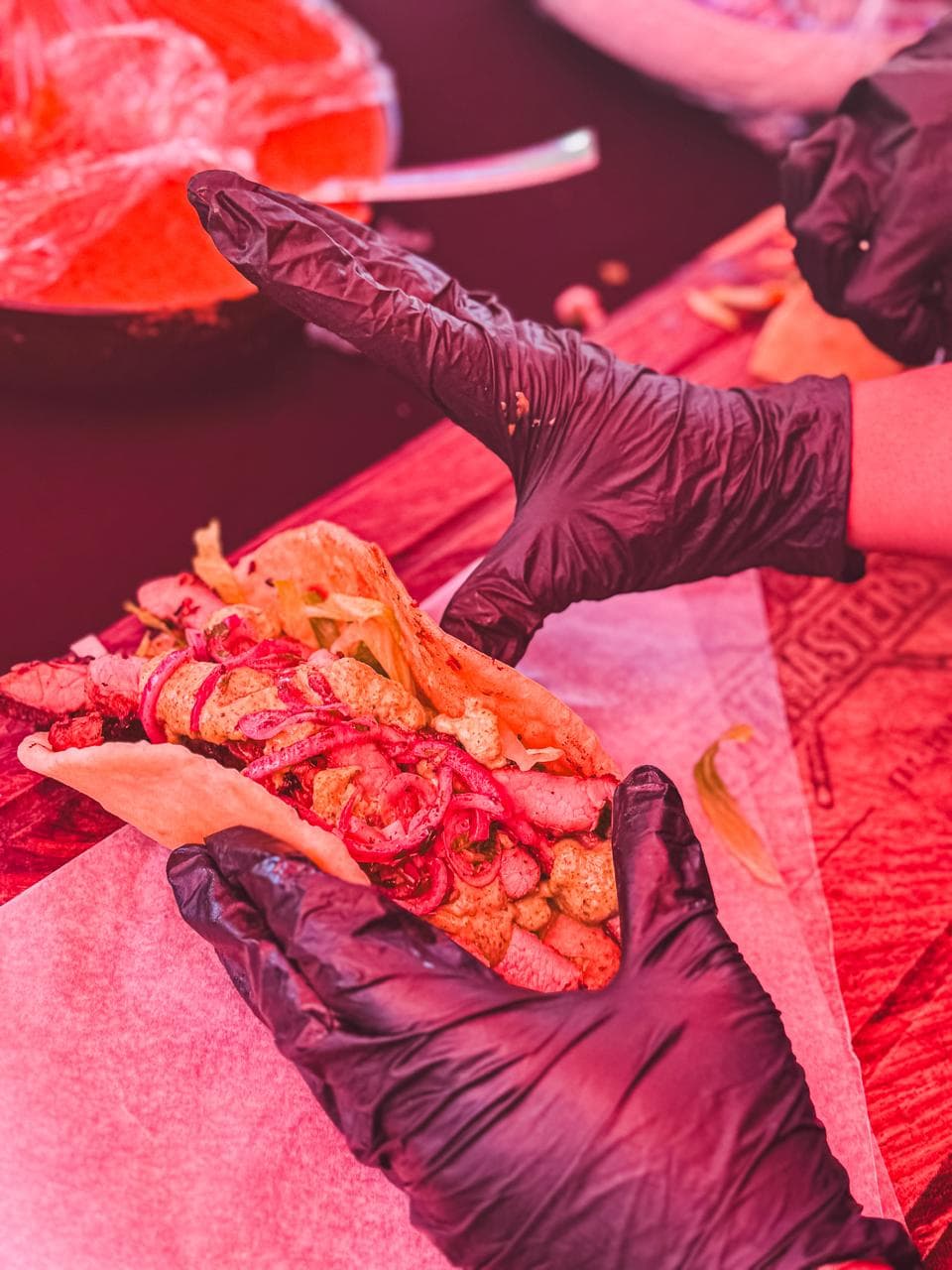 A grilled meat wrap folded with pickled red onion, held out under red festival lighting.