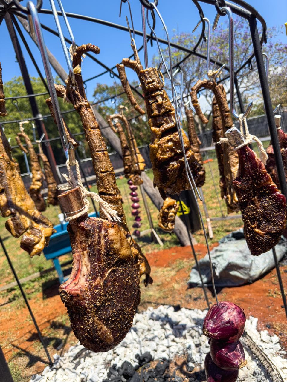 Cuts of meat hanging from a domed rig over glowing coals, red earth and a clear blue sky.