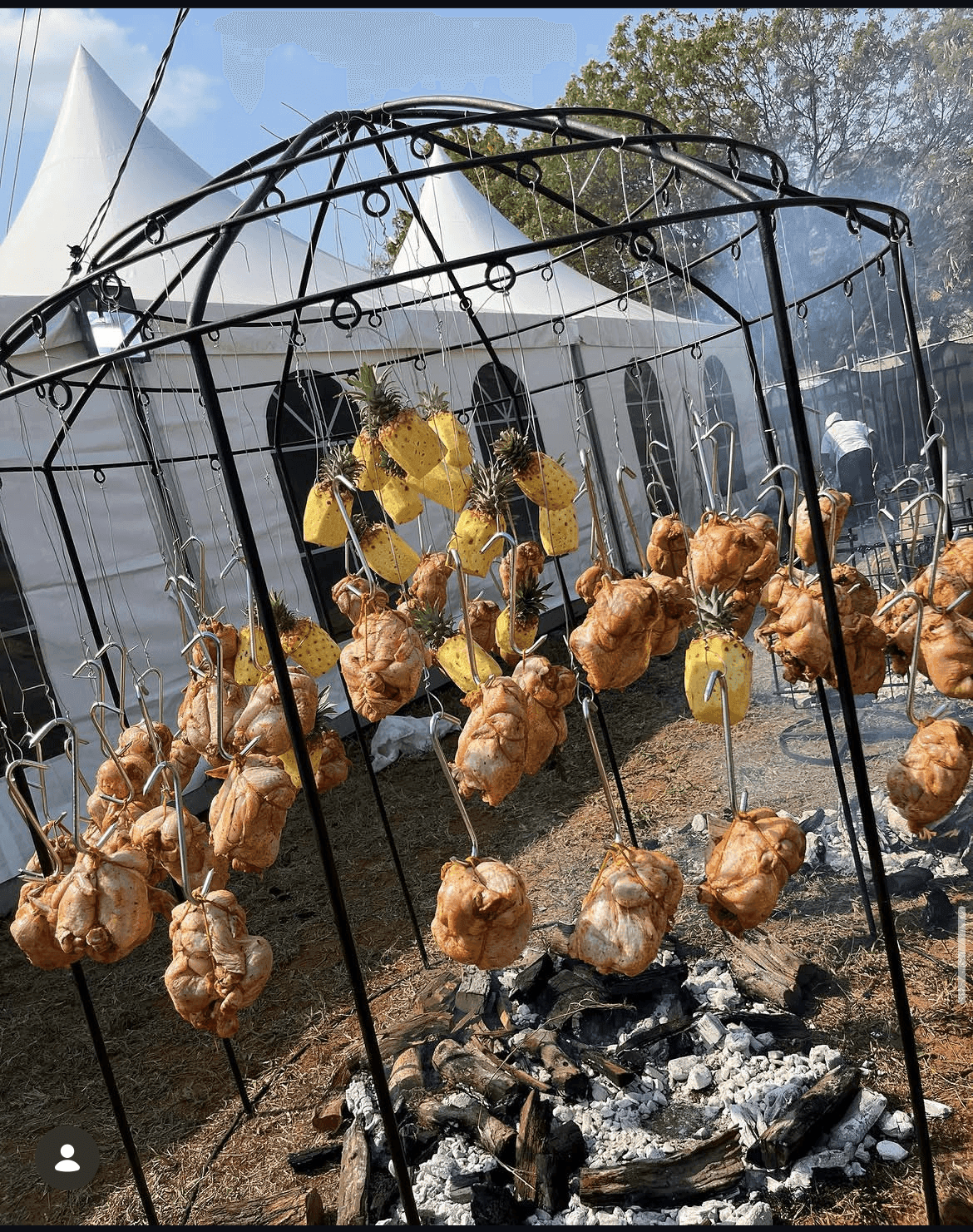 Whole chickens and pineapples hanging over glowing coals under a domed rig, festival marquee tents behind, at the Mozambique Barbecue Festival.