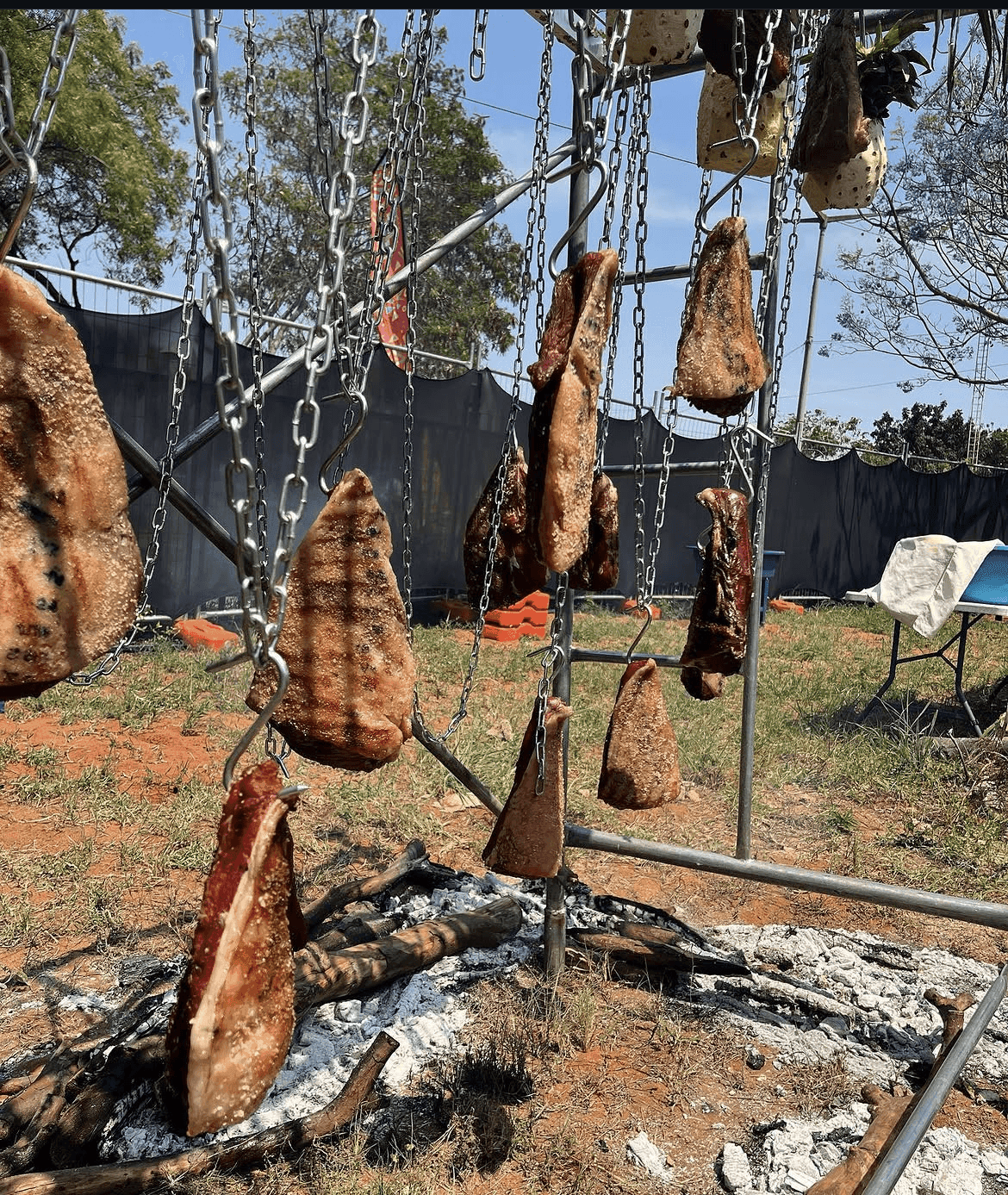 Whole cuts of meat hanging from chains over coals, black mesh fencing behind.