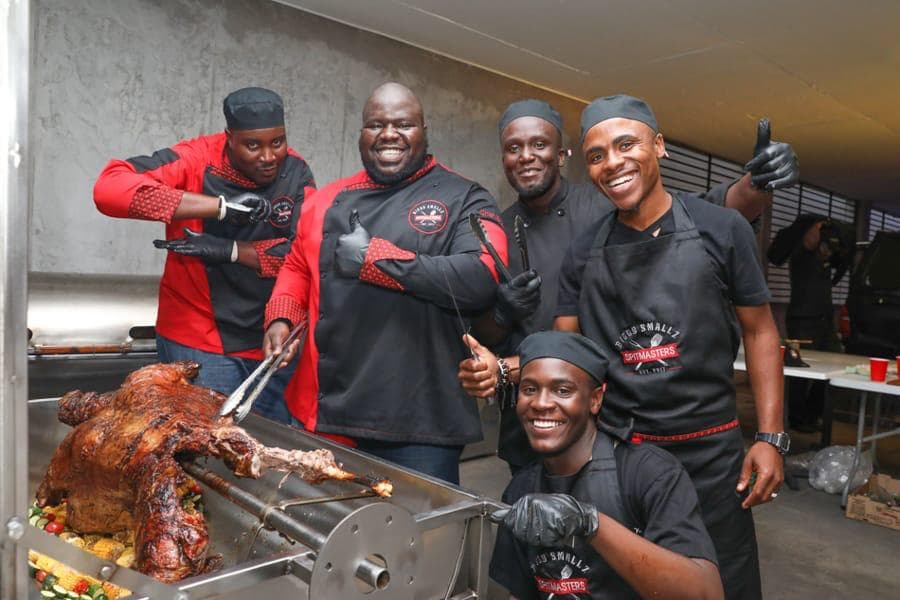 The full crew around a whole lamb on the spit, some in the earlier red-sleeve uniform.