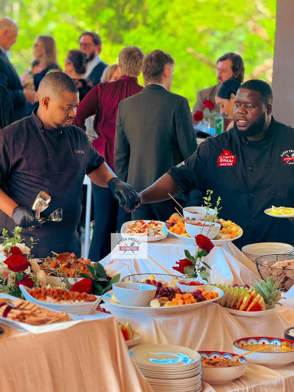 Biggy and Chef Sikolethu shaking hands over the harvest buffet, guests mingling behind them.